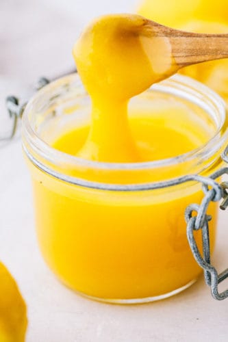 Lemon curd is a perfect combination of sweet and tangy with a smooth creamy texture. This lemony goodness comes together in a matter of minutes! Lemon curd in a small jar with a small wooden spoon with lemon curd dripping off the spoon.