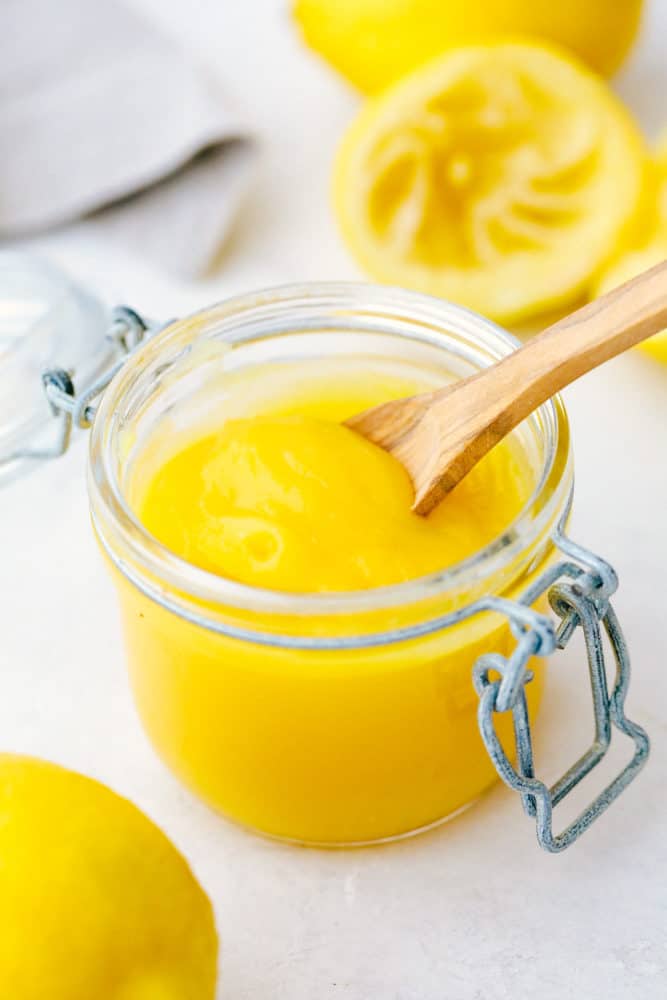Lemon curd is a perfect combination of sweet and tangy with a smooth creamy texture. This lemony goodness comes together in a matter of minutes! Lemon curd in an open jar with a small wooden spoon and lemons laying next to the jar as decoration.