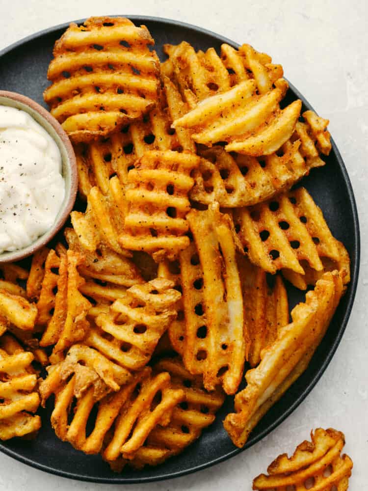 These air fryer frozen waffle fries are so easy and make the perfect side to any meal! Crispy and flavored to perfection, they will leave your mouth watering and wanting more! Waffle fries on a black plate with a small bowl of garlic aioli sauce.