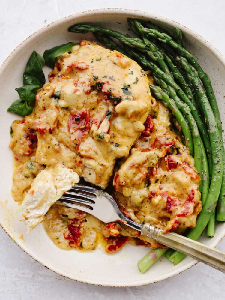 Marry me chicken is juicy and flavorful and smothered in a creamy parmesan sauce. Seasoned with savory herbs and sun-dried tomatoes, this dish will melt in your mouth, and will leave you begging for a proposal! A plate with chicken and asparagus. A silver fork is cutting into the chicken.