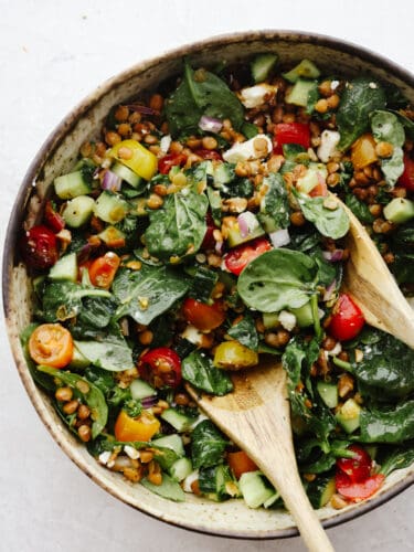 This hearty salad is a keeper! Filled with green lentils, spinach, cucumber, and crumbled feta cheese, drizzled with smooth Italian dressing, lentil salad is the best way to put a boost of flavor and nutrition into your day. Tossing lentil salad with 2 wooden spoons in a brown bowl.