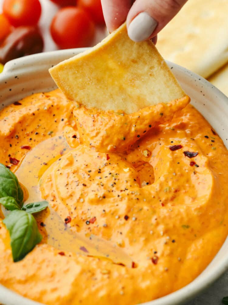 Roasted red pepper dip is smooth and creamy with the best warm spice! Serve it with your favorite chips for a boost of bold savory flavor! Closeup of a pita chip in red pepper dip.