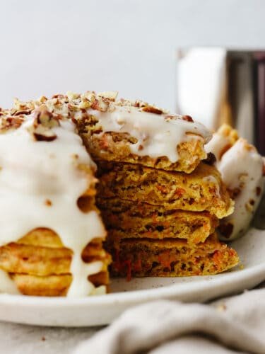 These carrot cake pancakes are fluffy, moist, and drizzled with a delicious cream cheese glaze that makes my mouth water! They will get gobbled up and fly off your table! A stack of carrot cake pancakes with a triangle bite cut out of them