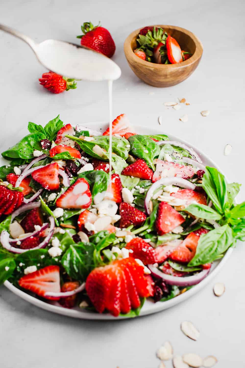Strawberry spinach salad is a perfect combination of fresh spinach, juicy strawberries, crunchy onions, and soft feta cheese; all tossed together with a creamy poppyseed dressing! All the flavor, texture, and deliciousness in one bowl! Poppyseed dressing being drizzled into a bowl of strawberry spinach salad.