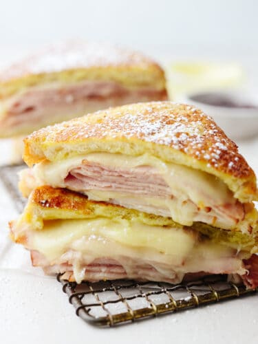 Get ready for the best Disneyland food out there with this copycat Monte Cristo sandwich!! The melted cheese combined with delicious turkey and ham sandwiched in crispy bread, all dipped into delicious raspberry jam... It's enough to make anyone's mouth water! A monte cristo sandwich, ready to eat on a cooling rack.