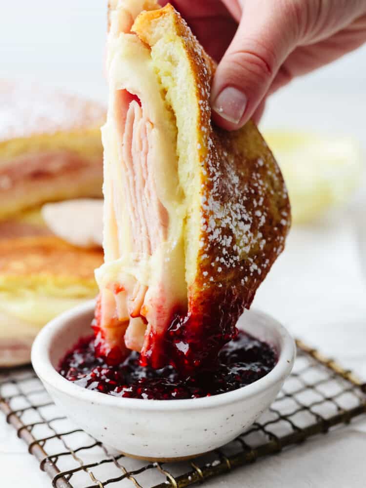Get ready for the best Disneyland food out there with this copycat Monte Cristo sandwich!! The melted cheese combined with delicious turkey and ham sandwiched in crispy bread, all dipped into delicious raspberry jam... It's enough to make anyone's mouth water! Half of a monte cristo sandwich being dipped into raspberry jam.