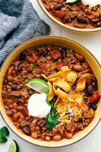 Thick, rich and flavorful! Filled with lean ground beef, mix of beans, ripe tomatoes, fresh seasonings and topped with all your favorites! This classic chili recipe is a MUST make! Chili in a bowl topped with lime wedge, sour cream, shredded cheese and Frito chips.