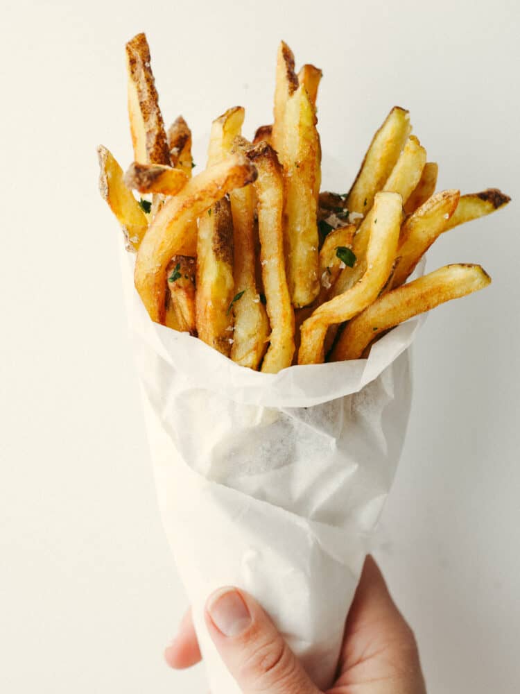 Try out these amazing Pommes Frites that are seasoned and cooked to perfection! They are the perfect side dish for just about anything. After all, everyone loves fries! Cooked Pommes Frites in a cone of parchment paper being held by a hand.