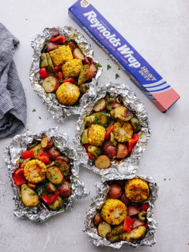 These foil pack grilled vegetables made with Reynolds Wrap® Heavy Foil are perfect for BBQ season! They cook just right on your grill and come out tender and seasoned to perfection. This is one side dish that you don't want to miss! 3 foild packs filled with cooked vegetables on a counter next to a box of Reynolds foil.