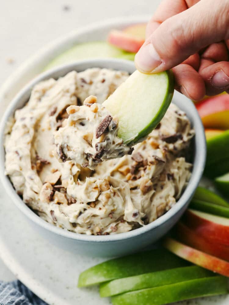 Toffee apple dip is a family favorite! A sweet dip complete with heath bar candy bits and cream cheese mixed with brown sugar, this dip is so delicious you won't be able to stop eating it! A close up of someone dipping an apple slice into apple toffee dip.