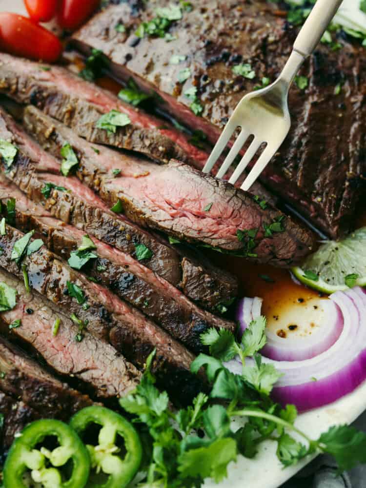 Enjoy this homemade carne asada recipe marinated in delicious Mexican flavors and spices. This juicy and tender flank steak is grilled to perfection for you to devour on its own or in any of your favorite Mexican dishes. Close up photo of a fork holding a slice of arne asada meat. Garnished with cilantro and onion and jalepeno on the side.