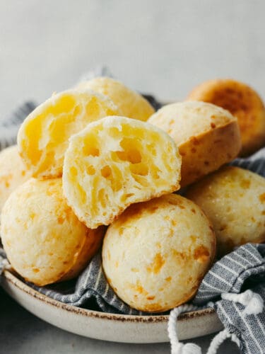 This delicious Brazilian cheese bread has a perfect combination of delicious taste and texture. Filled with pockets of airy goodness that taste so cheesy, you won't be able to stop at just one! A stack of Brazilian cheese bread with the top on broken in half so that you can see inside.