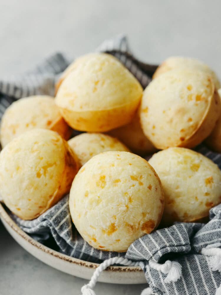 This delicious Brazilian cheese bread has a perfect combination of delicious taste and texture. Filled with pockets of airy goodness that taste so cheesy, you won't be able to stop at just one! A stack of Brazilian cheese bread on a plate with a kitchen towel.