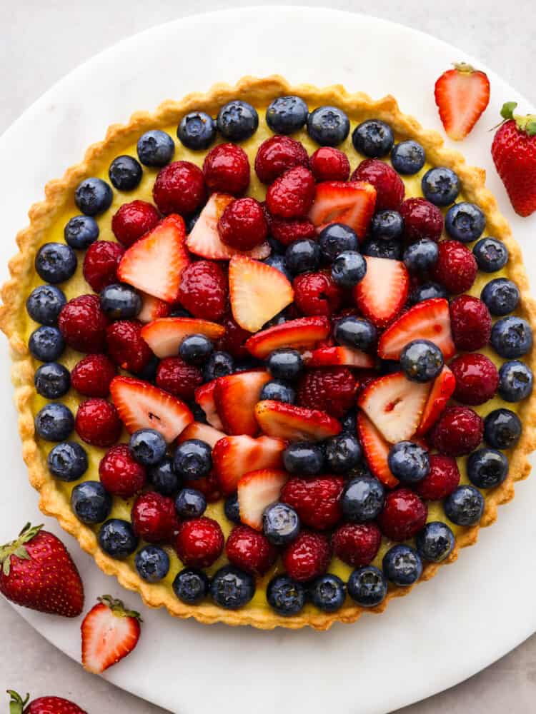 A buttery, flaky homemade crust, vanilla bean custard filling, and juicy berries make this berry tart the ultimate summer dessert! It's a beautiful and delicious way to showcase all the best fruit this season has to offer! Top-down view of a whole tart on a white plate.