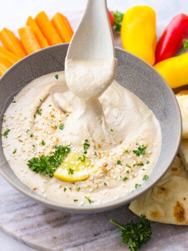 Get ready for the best tahini sauce that you have ever tasted! This makes any falafel, vegetable or salad taste better and it takes less than 5 minutes to put it all together! A bowl of tahini sauce on a platter with veggies. A plastic seerving spoon is scooping out some sauce.