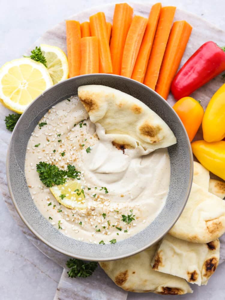 Get ready for the best tahini sauce that you have ever tasted! This makes any falafel, vegetable or salad taste better and it takes less than 5 minutes to put it all together! The top view of a bowl with tahini sauce garnished with parsley and sesame seds and a slice of pita bread being dipped into it.