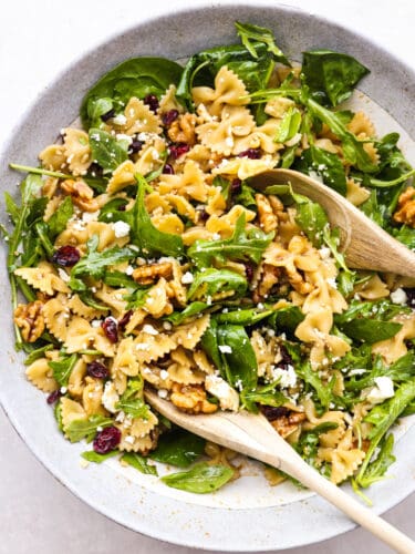 Cranberry feta bowtie salad with spinach is an easy salad perfect for potlucks and holidays! Each bite is packed with a zesty Dijon vinaigrette dressing, raisins, and walnuts. Top-down view of cranberry feta pasta salad being tossed with two wooden spoons.