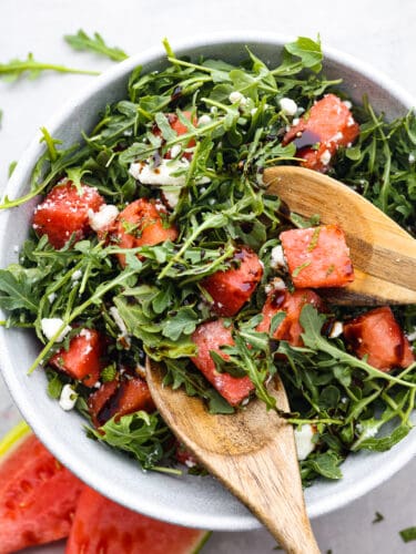 Nothing beats a fresh and healthy watermelon arugula salad in the summertime! The sweet watermelon is a surprising complement to the peppery arugula, creamy feta cheese, and balsamic glaze. Don't knock it until you try it! Top view of watermelon arugula salad in a large gray bowl. Wood serving spoons are sitting in the bowl of salad with watermelon and arugula scattered on the counter next to the bowl.