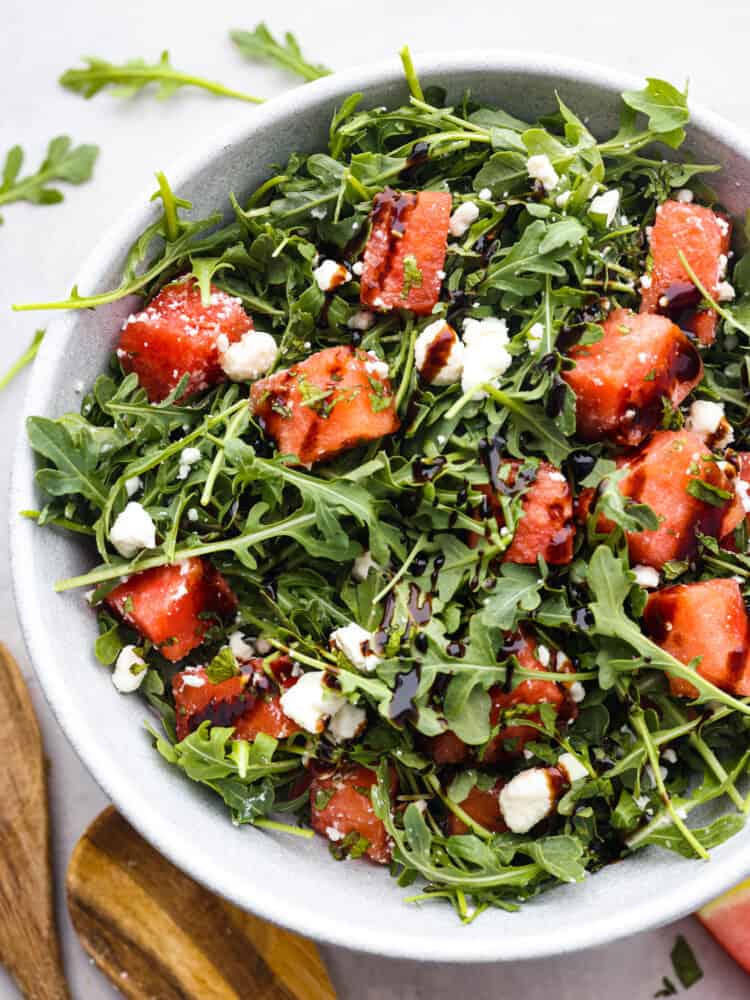 Nothing beats a fresh and healthy watermelon arugula salad in the summertime! The sweet watermelon is a surprising complement to the peppery arugula, creamy feta cheese, and balsamic glaze. Don't knock it until you try it! Top view of watermelon arugula salad in a gray bowl. Wood serving spoons and scattered arugula is laying next to the bowl.