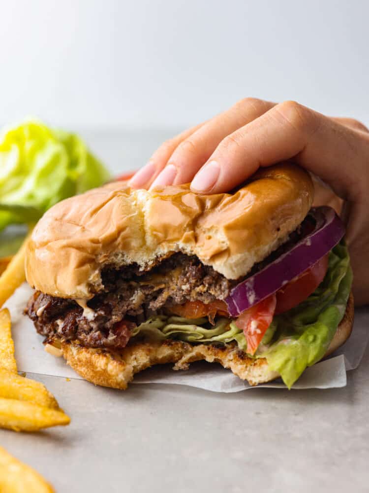 I love a good burger! Smash burgers are easy to prepare and come together so fast. If ever I'm craving a good old-fashioned burger, these smash burgers are my go-to recipe! Side view of a hand holding the smash burger with a bite taken out. Fries scattered on the side.