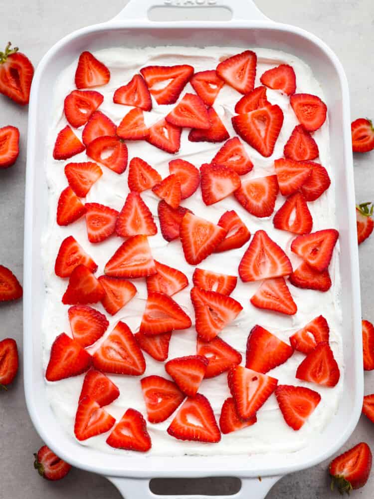 Incredibly light and tasty, this strawberry poke cake will be the star of any gathering! It's airy, soft, and brings that fresh strawberry taste into every single bite! The top view of a strawberry poke cake in the cake pan, topped with cool whip and sliced strawberries.