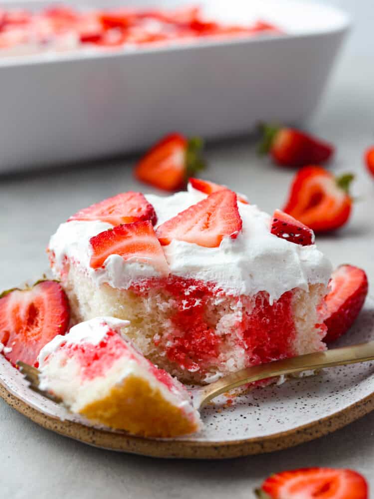 Incredibly light and tasty, this strawberry poke cake will be the star of any gathering! It's airy, soft, and brings that fresh strawberry taste into every single bite! A piece of strawberry poke cake on a plate with a gold fork cutting a bite.
