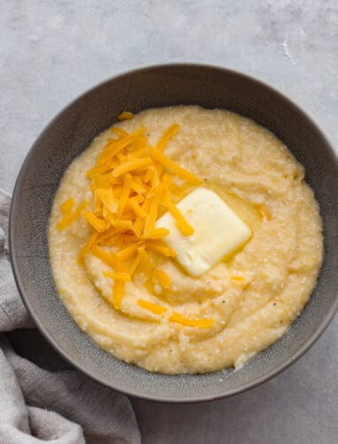 Creamy, cheesy, and comforting grits are a classic Southern dish. Slow-cooked cornmeal creates a deliciously smooth texture with a rich cheesy flavor. Top view of cheesy grits in a gray bowl. Garnished with melting butter and shredded cheddar cheese.