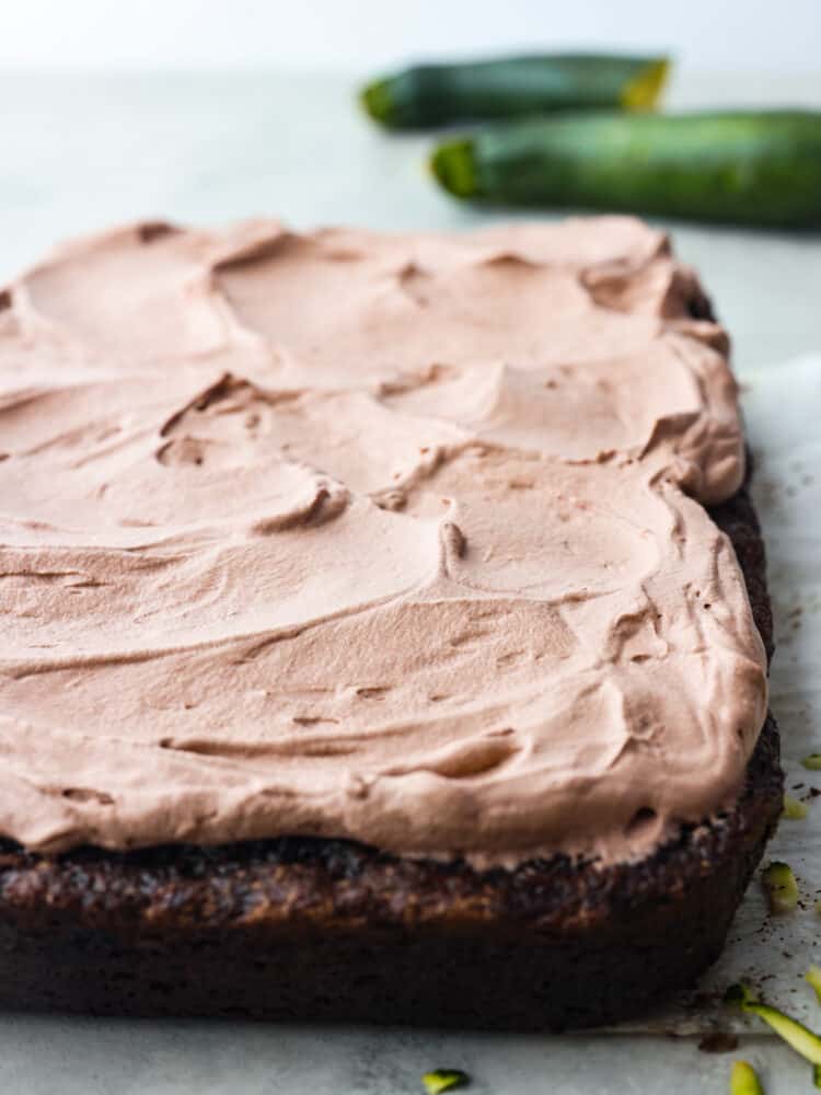 This Chocolate Zucchini Cake is a deep, dark chocolate cake that is topped with light and fluffy chocolate whipped cream! With 3 cups of zucchini, it is incredibly moist and you won't even notice all the vegetables! The side view of a chocolate zucchini cake on parchment paper with the whipped topping on top.