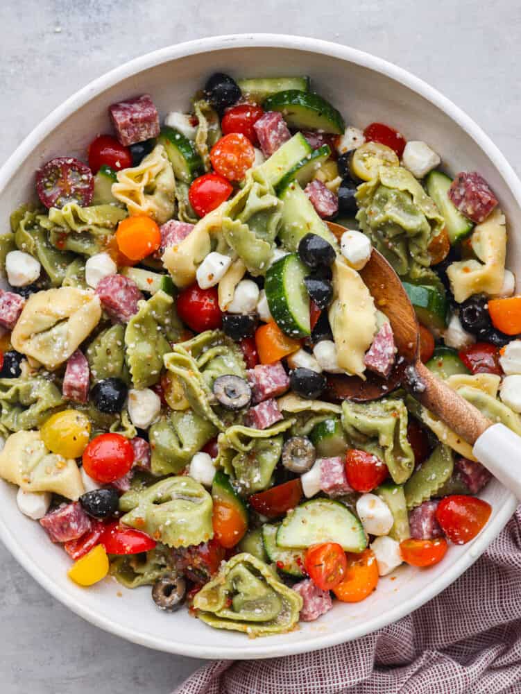 Easy Italian tortellini pasta salad is full of fresh summer veggies, salami, and tortellini pasta. It is tossed in a zesty Italian dressing and is always the hit of the potluck! Top view of tortellini pasta salad in a bowl with a wooden spoon.
