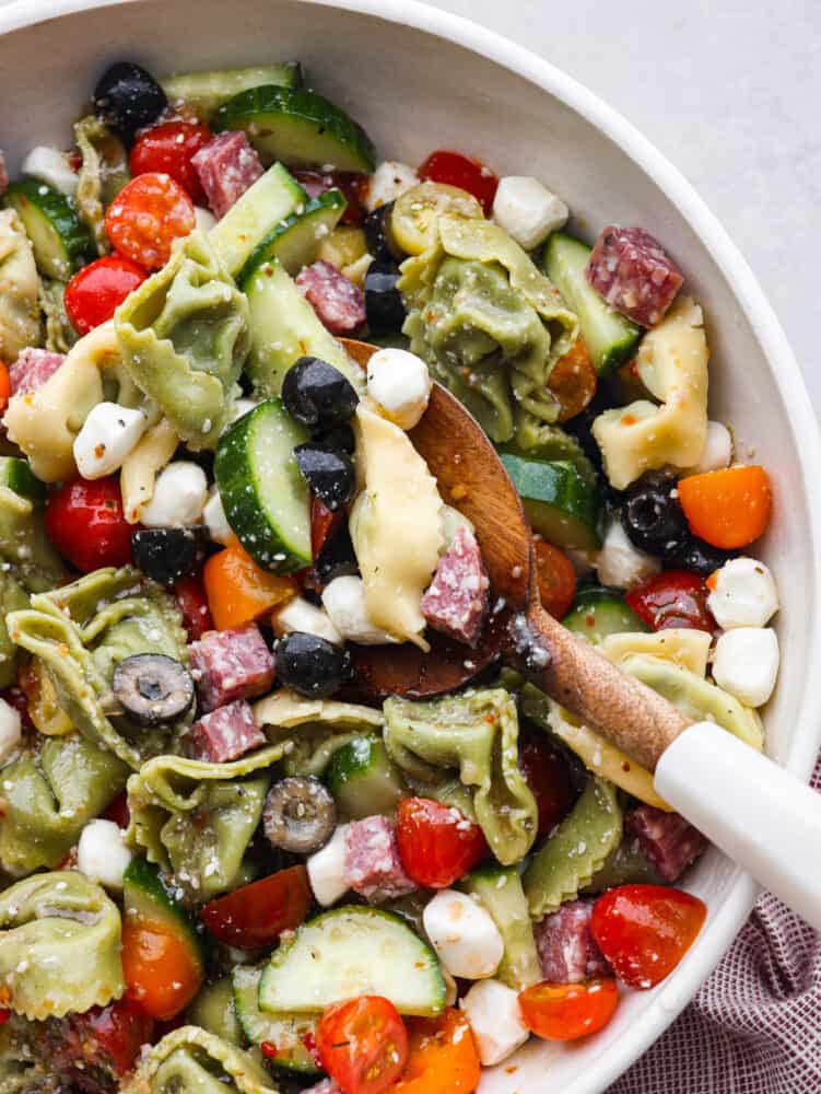 Easy Italian tortellini pasta salad is full of fresh summer veggies, salami, and tortellini pasta. It is tossed in a zesty Italian dressing and is always the hit of the potluck! A bowl of Italian tortellini pasta salad with a wooden spoon.