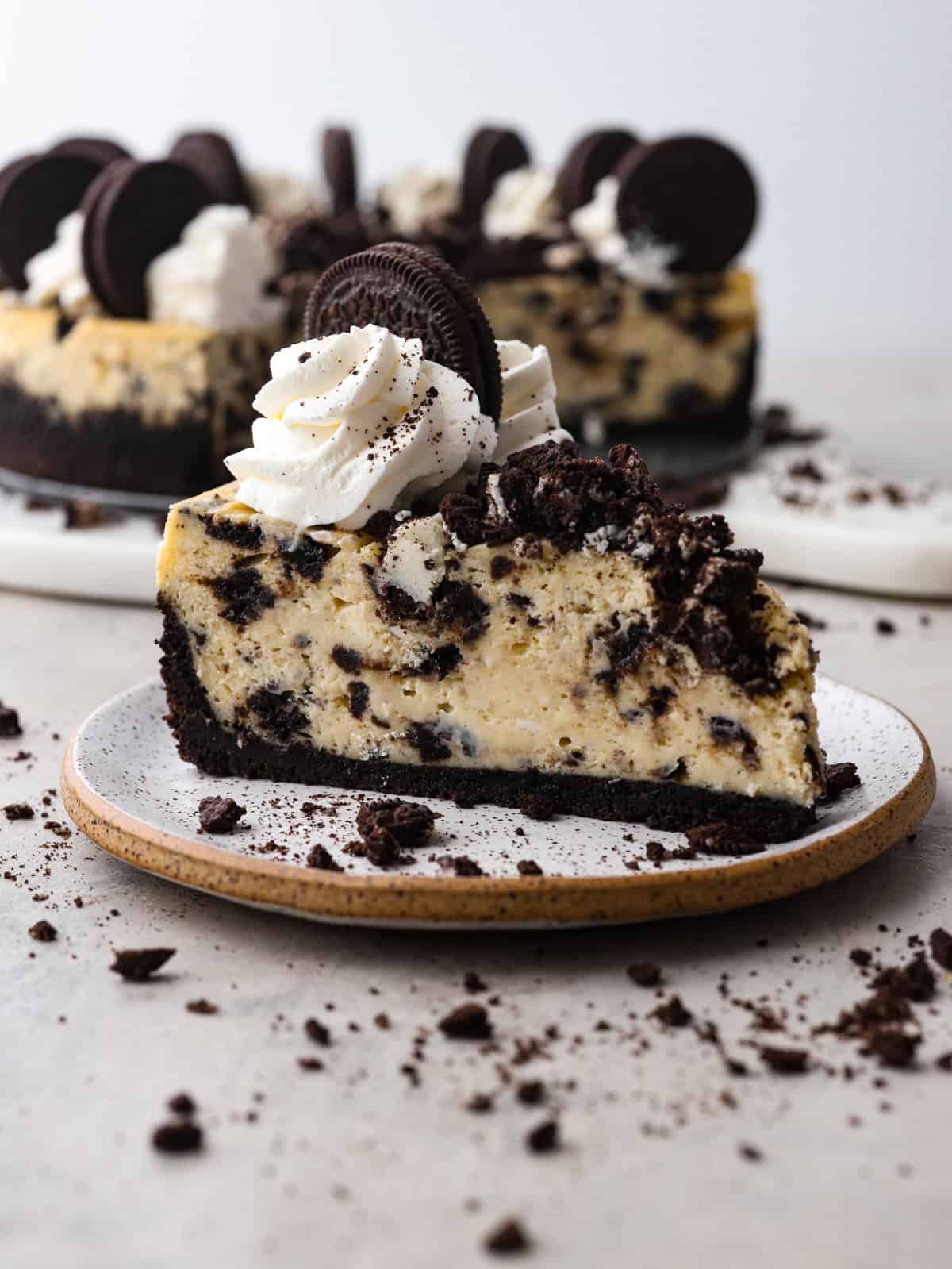 Let your teeth sink into the most delectable, creamy oreo cheesecake that you have ever had! Silky and smooth with the perfect bite-sized oreo bits in every single slice, you won't be able to stop raving about this cheesecake! A slice of oreo cheescake on a plate with some oreo crumbs sprinkles around it.